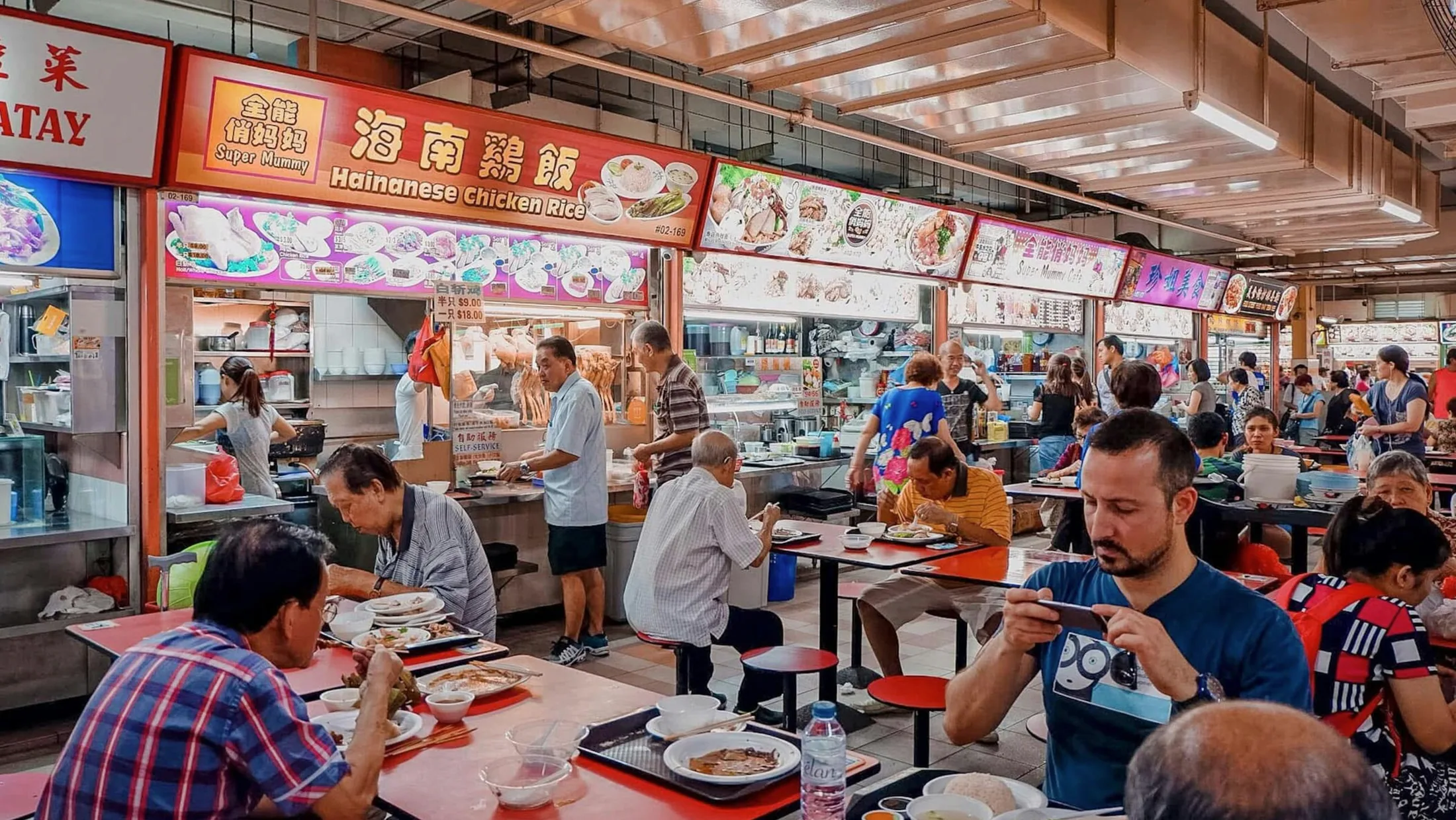 A busy Singapore hawker centre with bright food stalls and diners at red tables.