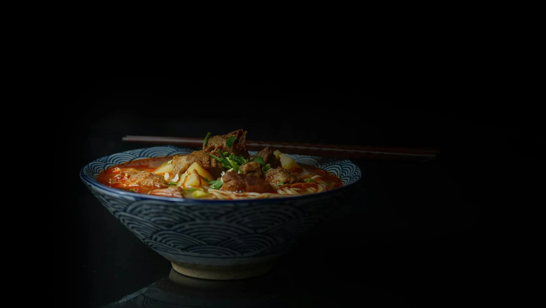 A dark cinematic bowl of hot soup and pork ribs on a black table.